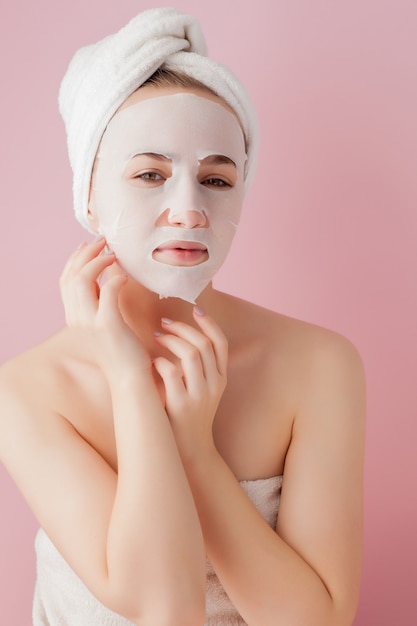 De mooie jonge vrouw past een kosmetisch weefselmasker toe op een gezicht op een roze ruimte