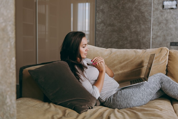 De mooie jonge vrouw thuis liggend op de bank, ontspannen in haar woonkamer en werken met laptop