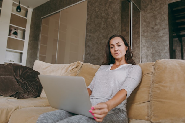 De mooie jonge vrouw thuis zittend op de bank, ontspannen in haar woonkamer en werken met laptop