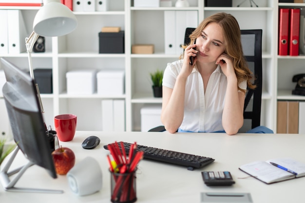 De mooie jonge vrouw zit in het kantoor en spreekt aan de telefoon.