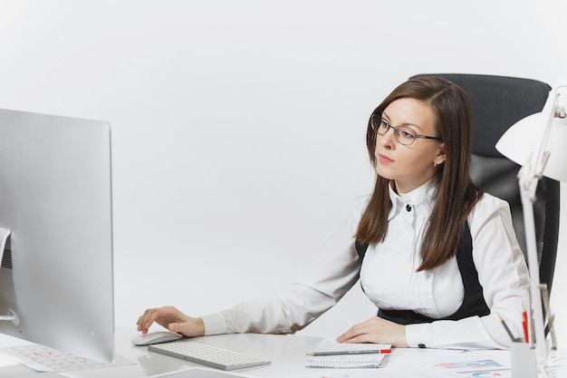 De mooie serieuze en verdiepte bruinharige zakenvrouw in pak en bril zit aan het bureau, werkend op de computer met moderne monitor met documenten in een licht kantoor,