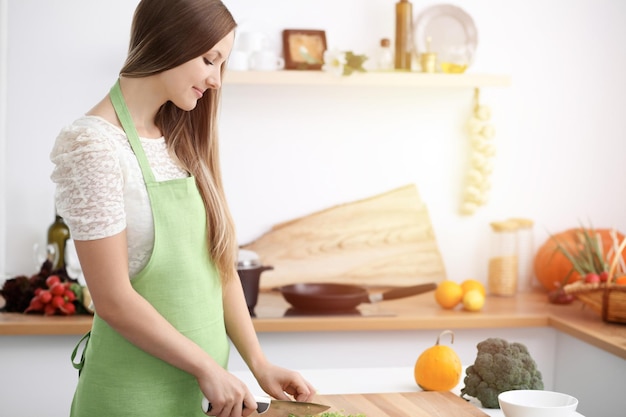 De mooie vrouw gekleed in schort kookt maaltijd in zonnige keuken.