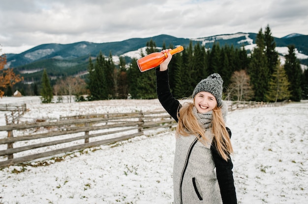 De mooie vrouw houdt een flessenchampagne tegen de achtergrond van de winterbergen. Meisje in de besneeuwde winter, wandelen in de natuur. Reis- en vakantieconcept. Vakantie winterseizoen.