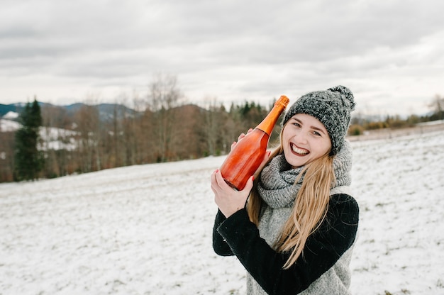 De mooie vrouw houdt een flessenchampagne tegen de de winterbergen. Meisje in de besneeuwde winter, wandelen in de natuur.