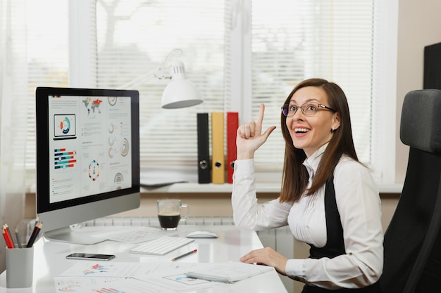 De mooie zakenvrouw in pak en bril die op de computer werkt met documenten in een licht kantoor, omhoog kijkend