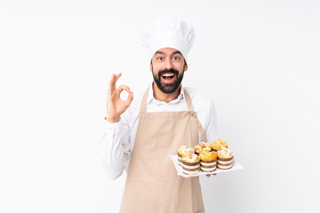 De muffincake van de jonge mensenholding over geïsoleerd wit verrast en tonend ok teken