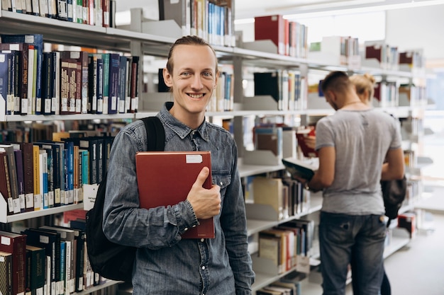 De nadenkende jongen kijkt als een student die zich met boek in de bibliotheek van een universiteit bevindt