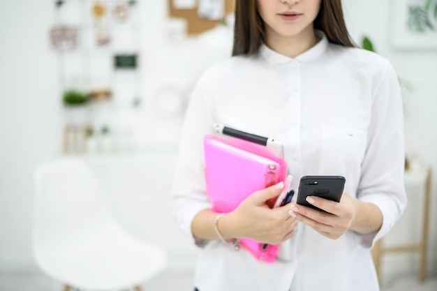 De officemanager houdt een roze map met documenten en een digitale tablet vast Vrouw secretaresse typt op mobiel Meisje in een wit overhemd op het werk