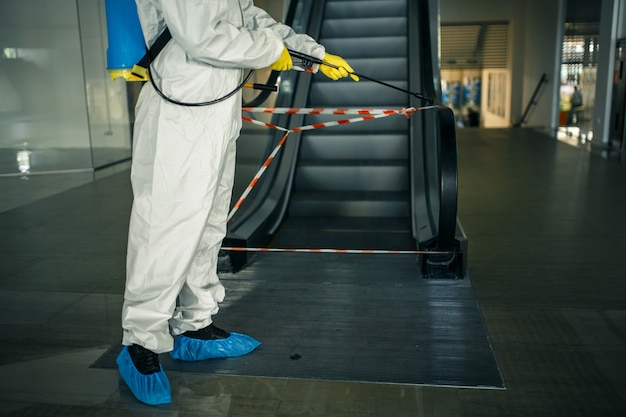 De ontsmettingsmedewerker desinfecteert de roltrap met een spray in het lege winkelcentrum om te voorkomen dat de covid-19 zich op openbare plaatsen verspreidt.