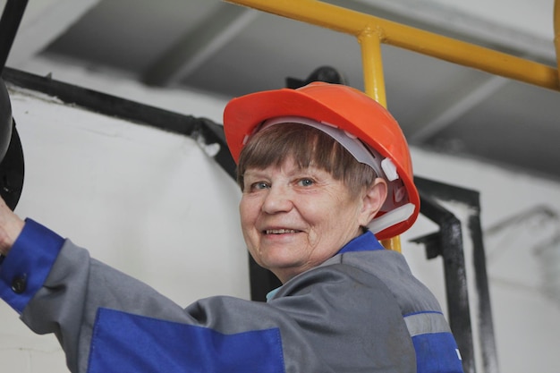 De oudere vrouw in werkoverall en rode helm op de productielocatie