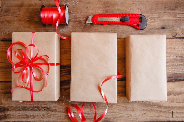 De platte lay-out van ambachtelijke dozen, rood lint en rood mes op houten tafel. Verpakken van geschenken proces.