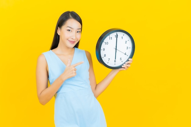 De portret mooie jonge aziatische vrouw toont wekker of klok op gele kleurenmuur