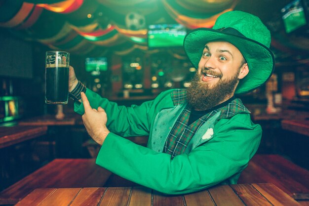 De positieve en vrolijke jonge mens in groen kostuum zit bij lijst in bar. Hij wijst op mok met donker bier. Guy ziet er blij uit.