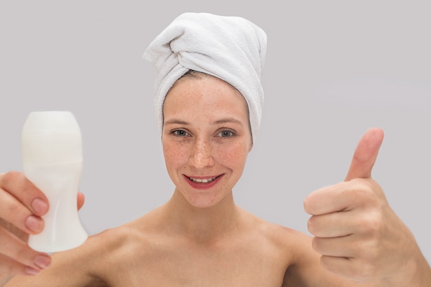 De positieve en vrolijke jonge vrouw bevindt zich en kijkt. ze houdt haar grote duim omhoog en deodorant in een andere hand. er is een witte handdoek om haar haar.