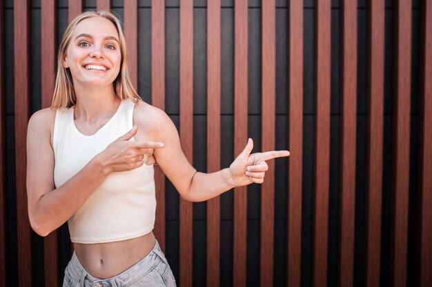 De positieve jonge vrouw in wit overhemd bevindt zich en stelt. Ze glimlacht en wijst naar rechts.