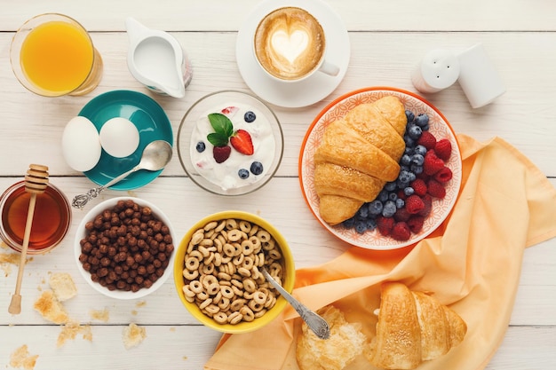 De rijke achtergrond van het ontbijtmenu. Franse knapperige croissants, muesli, glas verse sinaasappel, yoghurt, koffie, bessen, honing en gekookte eieren voor smakelijke ochtendmaaltijden op houten tafel, bovenaanzicht