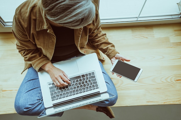 De rijpe vrouw bekijkt het zwarte scherm van mobiele telefoon die met laptop computer werken