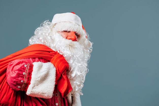 De rode neus van de kerstman op het blauwe vooraanzicht als achtergrond leunt onder het gewicht van een rode tas met