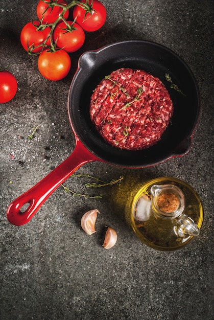 De ruwe organische koteletten van rundvleesvleesburgers met kruiden, tijm, tomaten, olijfolie in pan op zwarte, hoogste mening