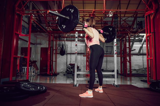 De schitterende jonge geschiktheidsvrouw buigt en doet ploegoefening met een bar achter de hals in de gymnastiek.