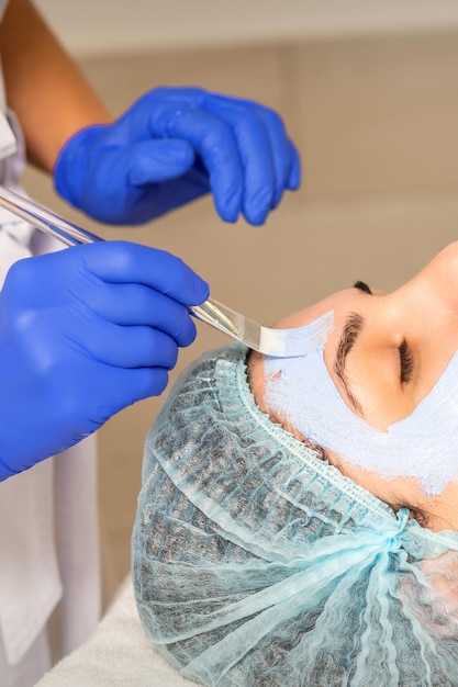 De schoonheidsspecialiste met borstel brengt een fotochemisch en glycolisch peeling-gezichtsmasker aan op het gezicht van de vrouwelijke patiënt in de schoonheidskliniek