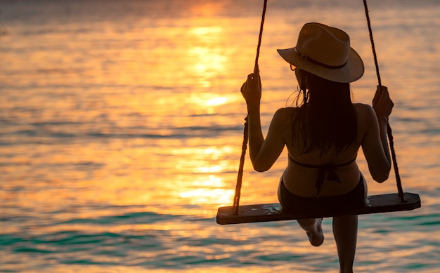 De silhouetvrouw draagt bikini en de strohoed slingert de schommels bij het strand op de zomervakantie bij zonsondergang