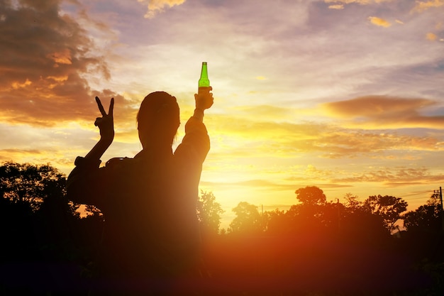 De silhouetvrouw hief handen op houdend een groene bierfles op de zonsonderganghemel,