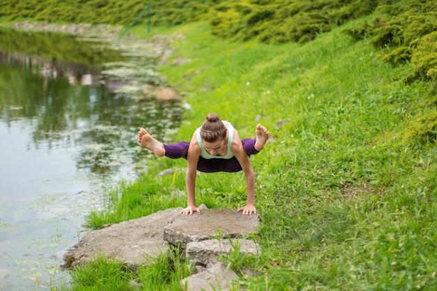 Foto de slanke jonge donkerbruine yogi voert complexe yogaoefening op groen gras in de zomer op een achtergrond van aard uit