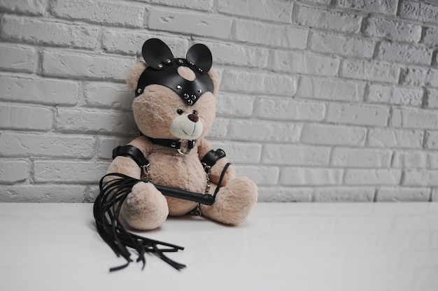 De speelgoedteddybeer gekleed in leren riemen en maskeraccessoire voor BDSM-spellen op een lichte achtergrondstructuur van een bakstenen muur