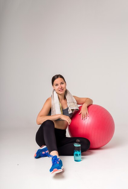 De sportvrouw zit met een geschiktheidsbal en water en een handdoek op een wit en glimlacht