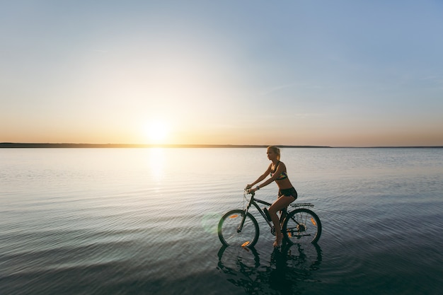 De sterke blonde vrouw in een kleurrijk pak zit op de fiets in het water bij zonsondergang op een warme zomerdag. Geschiktheidsconcept.