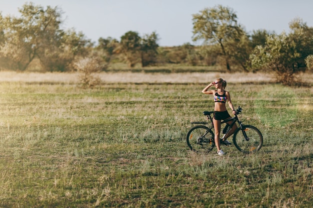 De sterke blonde vrouw in een kleurrijk pak zit op een fiets in een woestijngebied met bomen en groen gras en kijkt naar de zon. Geschiktheidsconcept.