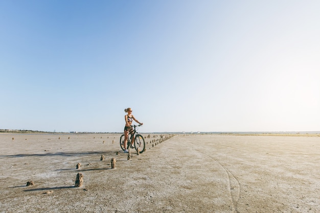 De sterke blonde vrouw in een veelkleurig pak en zonnebril zit op een fiets in een woestijngebied en kijkt naar de zon. Geschiktheidsconcept.