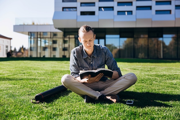 De student leest een boek zittend op het groene gazon vóór de moderne bibliotheek