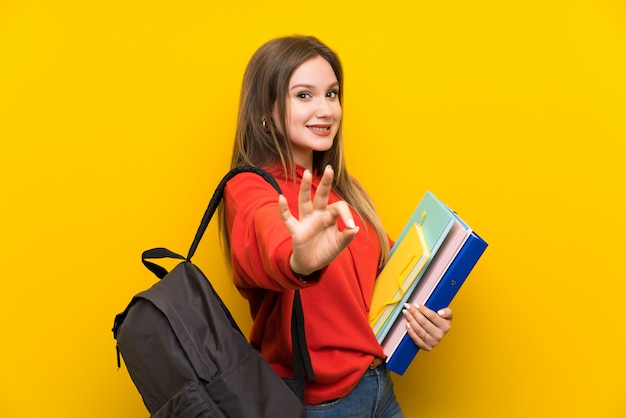 De studentenmeisje van de tiener over geel tonend een ok teken met vingers