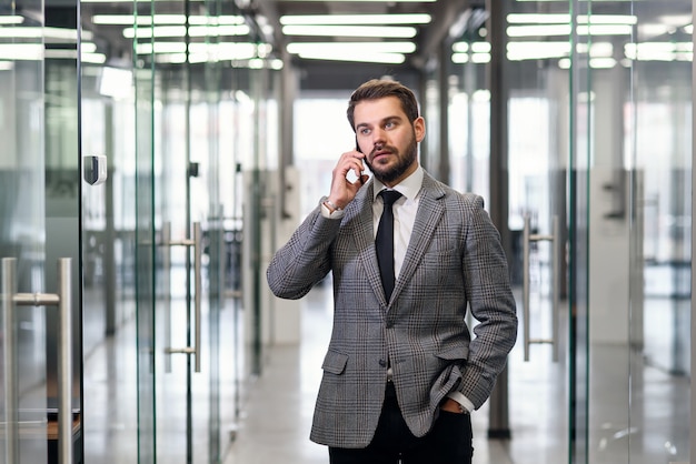 De succesvolle bedrijfsmens heeft telefoongesprek terwijl status in lege bureaugang. Mannelijke professionele bankier in pak praten op mobiele telefoon tijdens werkpauze.