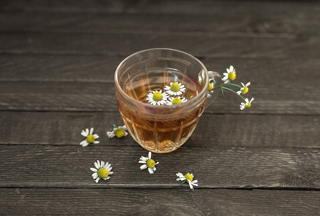 De theekop van het glas met kamille op een houten achtergrond. Kamillebloemen zijn overal verspreid.