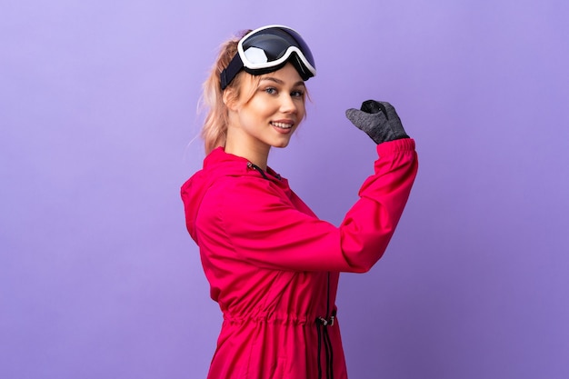 De tienermeisje van de skiër met snowboardglazen over geïsoleerde purpere muur die sterk gebaar doet