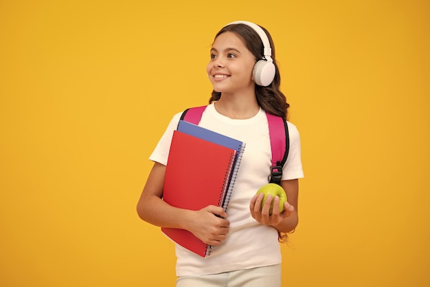 De tienerstudent van het schoolmeisje in hoofdtelefoons houdt boeken op gele geïsoleerde studioachtergrond School