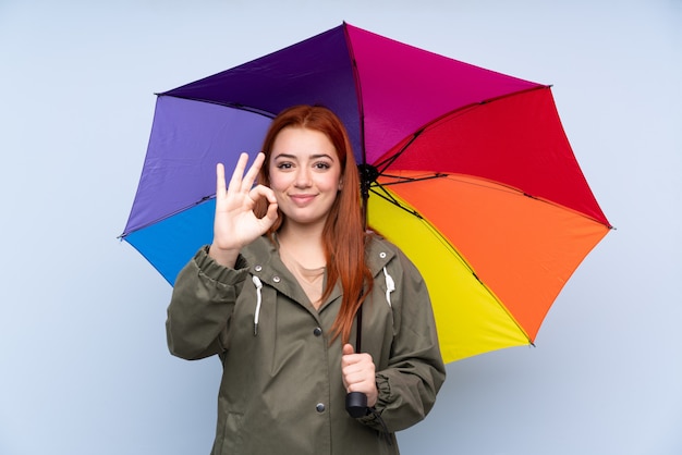 De tienervrouw die van de roodharige een paraplu over geïsoleerde blauwe muur houdt die een ok teken met vingers toont