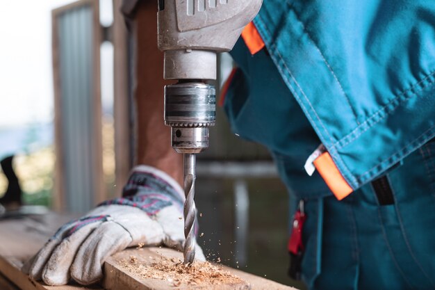 De timmerman in overall en handschoenen boort een houten raad in de huisworkshop