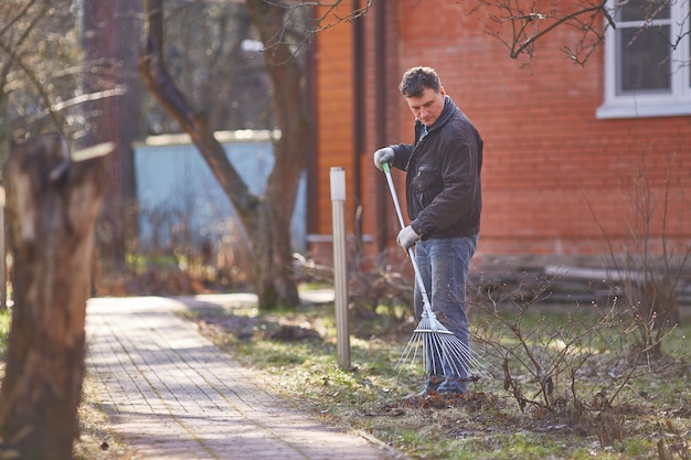 De tuinman harkt herfstbladeren in de tuin van een huisje van rode baksteen.