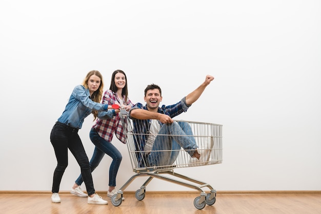 De twee vrouwen en een man plezier met een winkelwagentje op een witte muur achtergrond