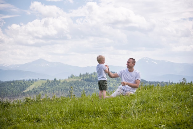 De vader en de jonge zoon blazen paardebloemen die in het gras op een van groen bos, bergen en hemel met wolken zitten. Vriendschap