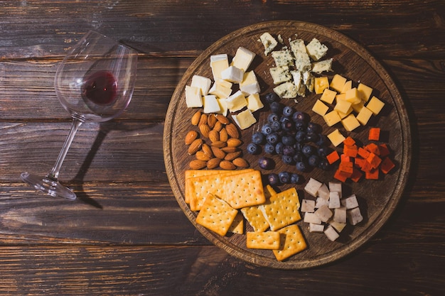 De verschillende soorten kaas en andere snacks liggen op een houten dienblad. Naast de houten tafel staat een glas met een druppel rode wijn