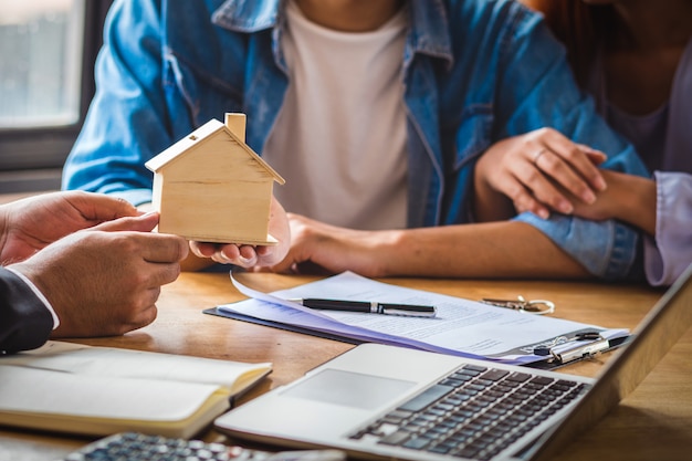 De vertegenwoordiger van de close-upverkoop biedt het mockup van het houten huis aan Aziatisch jong paar aan