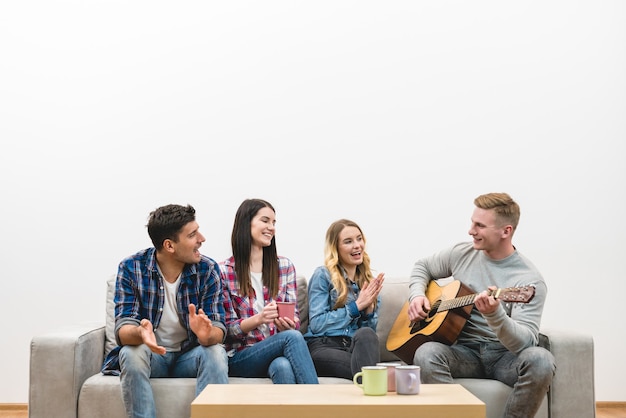 De vier gelukkige mensen met een gitaar zitten op de bank op de witte muurachtergrond