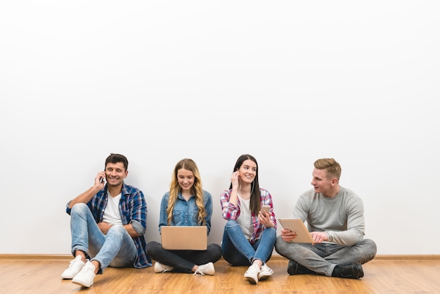 De vier mensen met gadgets zitten op de vloer op de witte muurachtergrond