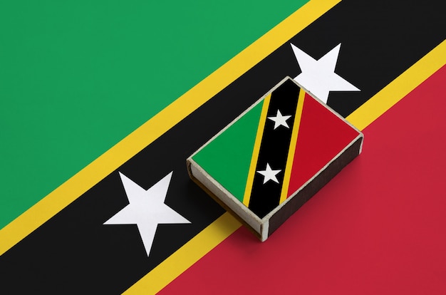 De vlag van Saint Kitts en Nevis staat afgebeeld op een luciferdoosje dat op een grote vlag ligt