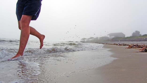 De voeten van een man die bij mistig weer langs een zeestrand rent Spatten van de golven vliegen rond zand en golven onder de voeten van een man die in korte broek rent Barefoot ochtend joggen op het strand